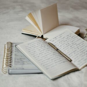 Close-up of open notebooks and a pen on a soft bed surface with delicate dried flowers.